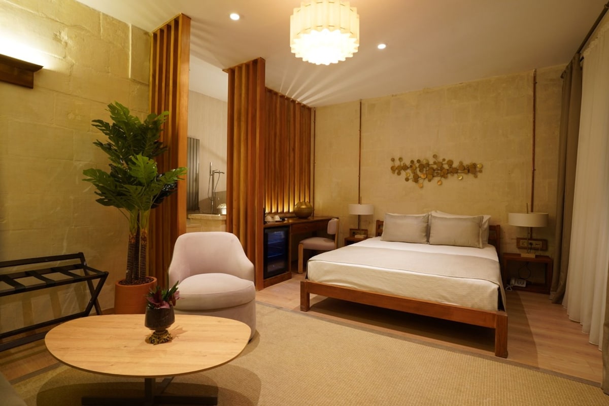 A hotel room with a bed, a desk, a chair, a small table, and a potted plant.