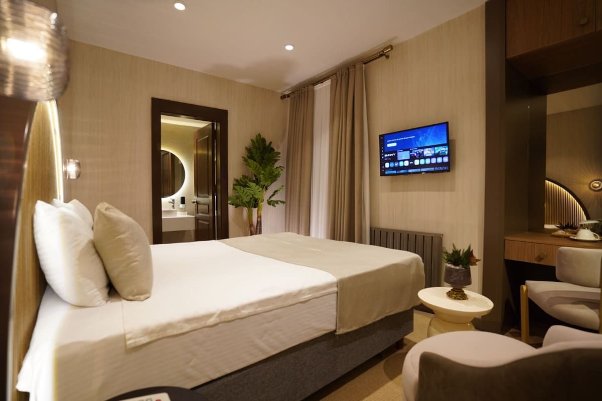 Habitación de hotel con una cama grande, un televisor montado en la pared, un escritorio con una silla y una mesa redonda con una lámpara.