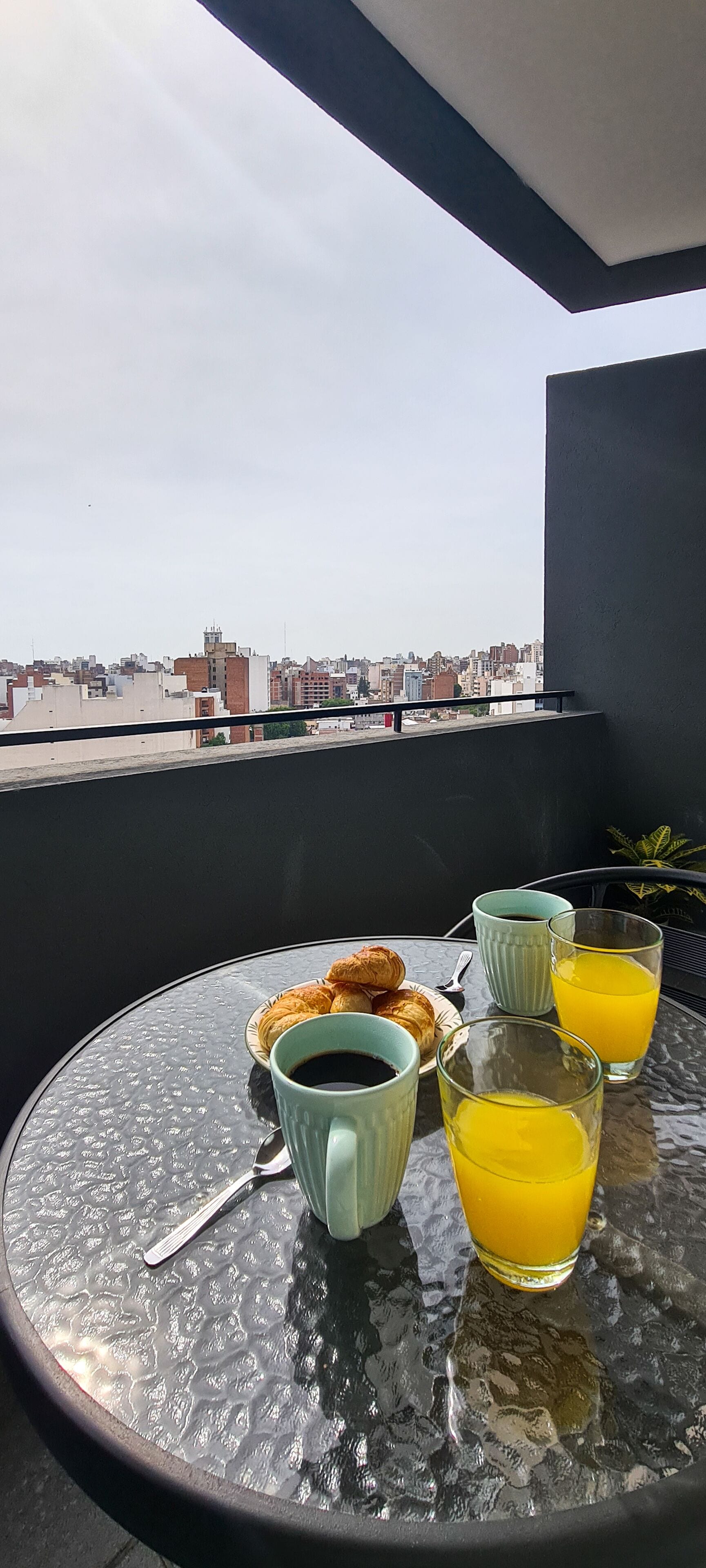 A table with croissants, coffee, and orange juice on a balcony overlooking a cityscape.