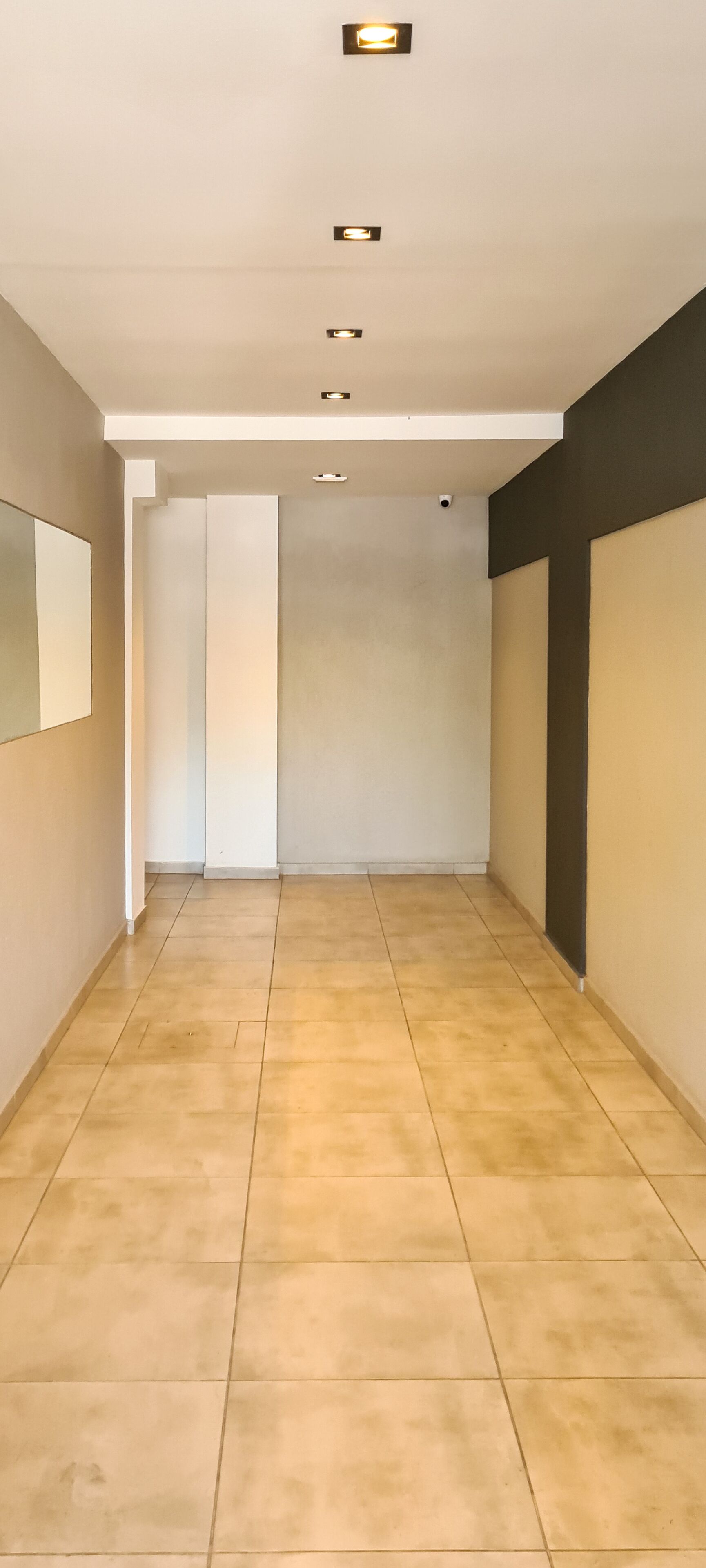 A hallway with a tiled floor, recessed lighting, and a plain wall with a mirror.
