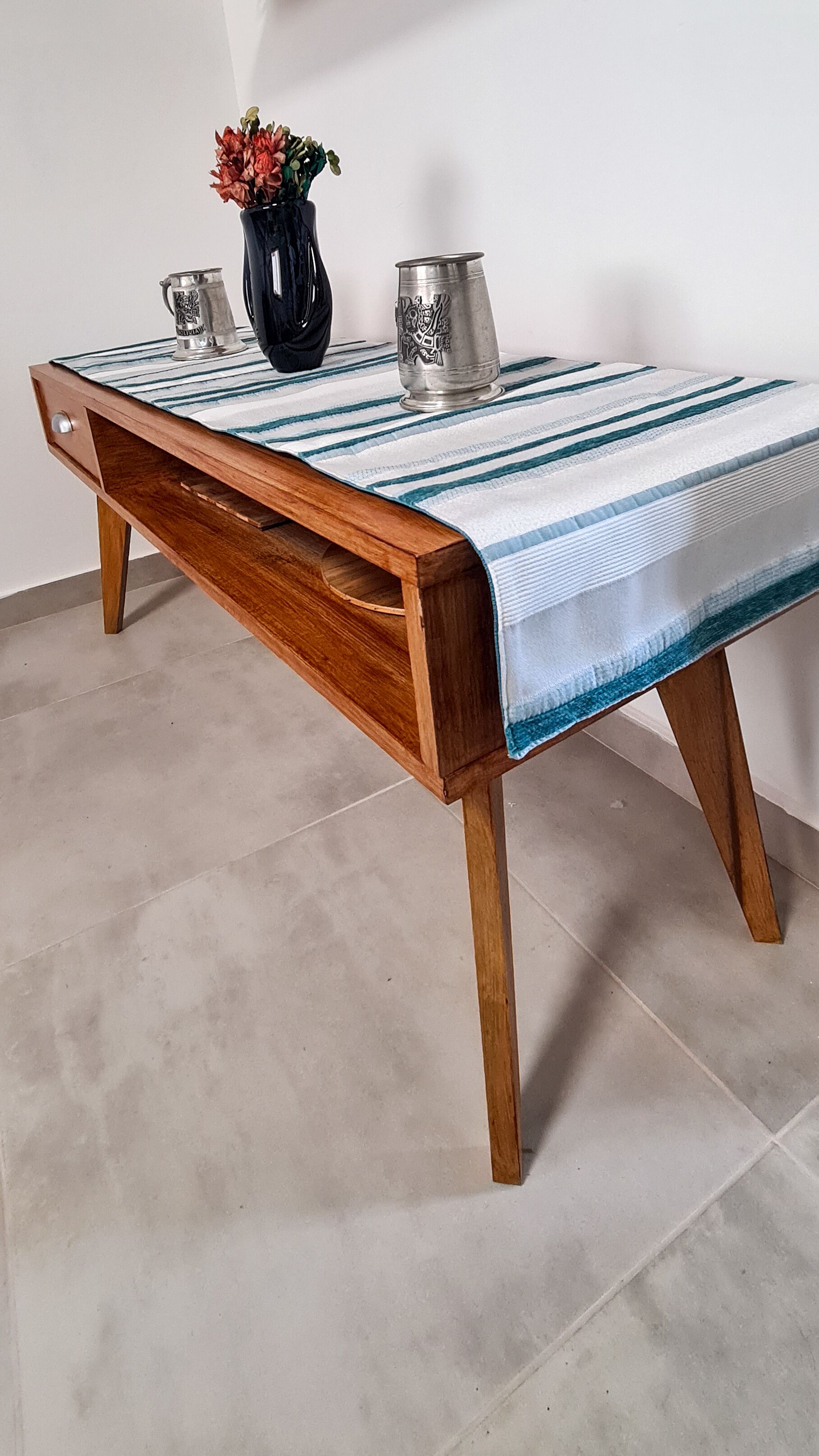 A wooden table with a striped tablecloth, two metal pitchers, and a vase with flowers.