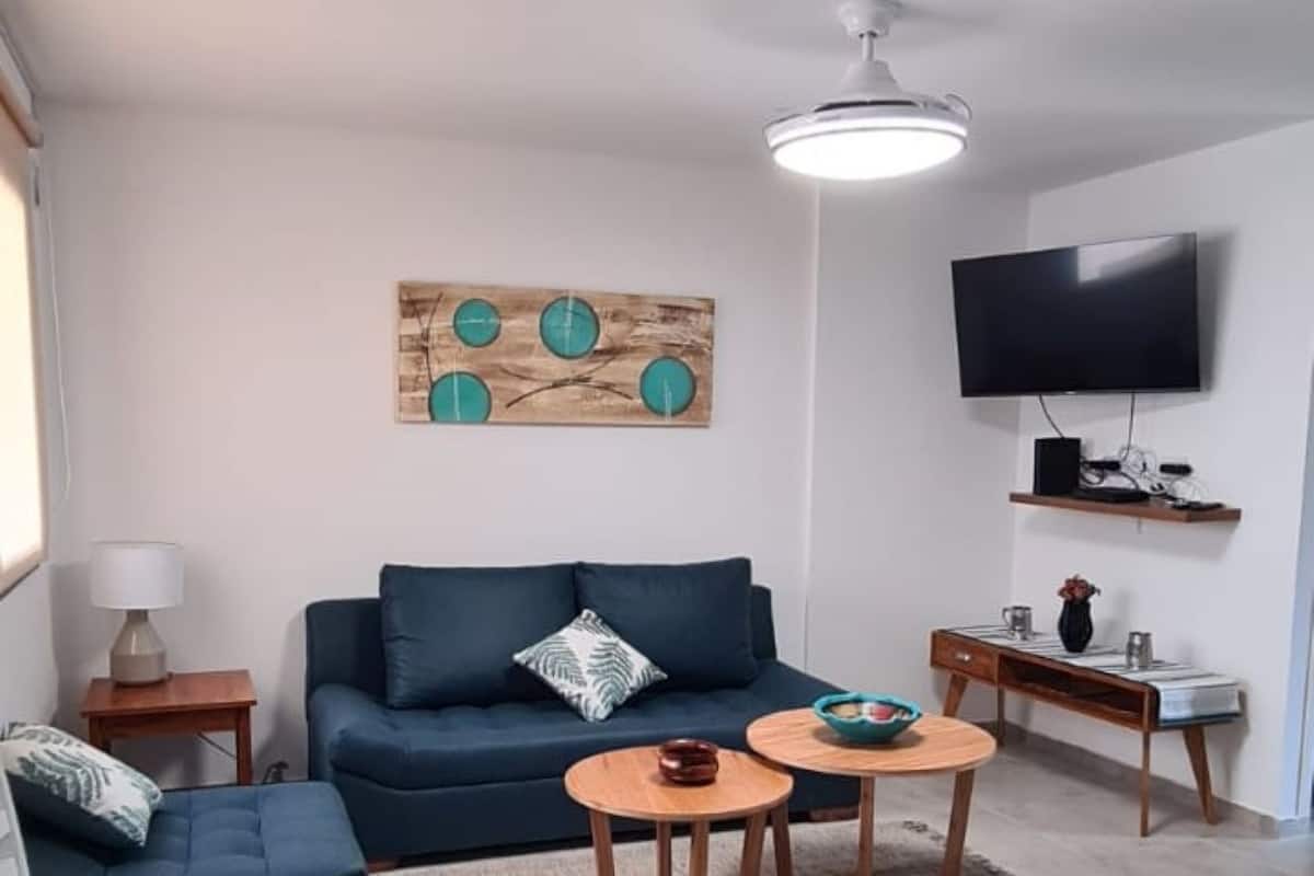 Uma sala de estar moderna com um sofá azul escuro, duas mesas redondas de madeira, uma pequena mesinha lateral, uma TV de tela plana fixada na parede e um objeto decorativo na parede.