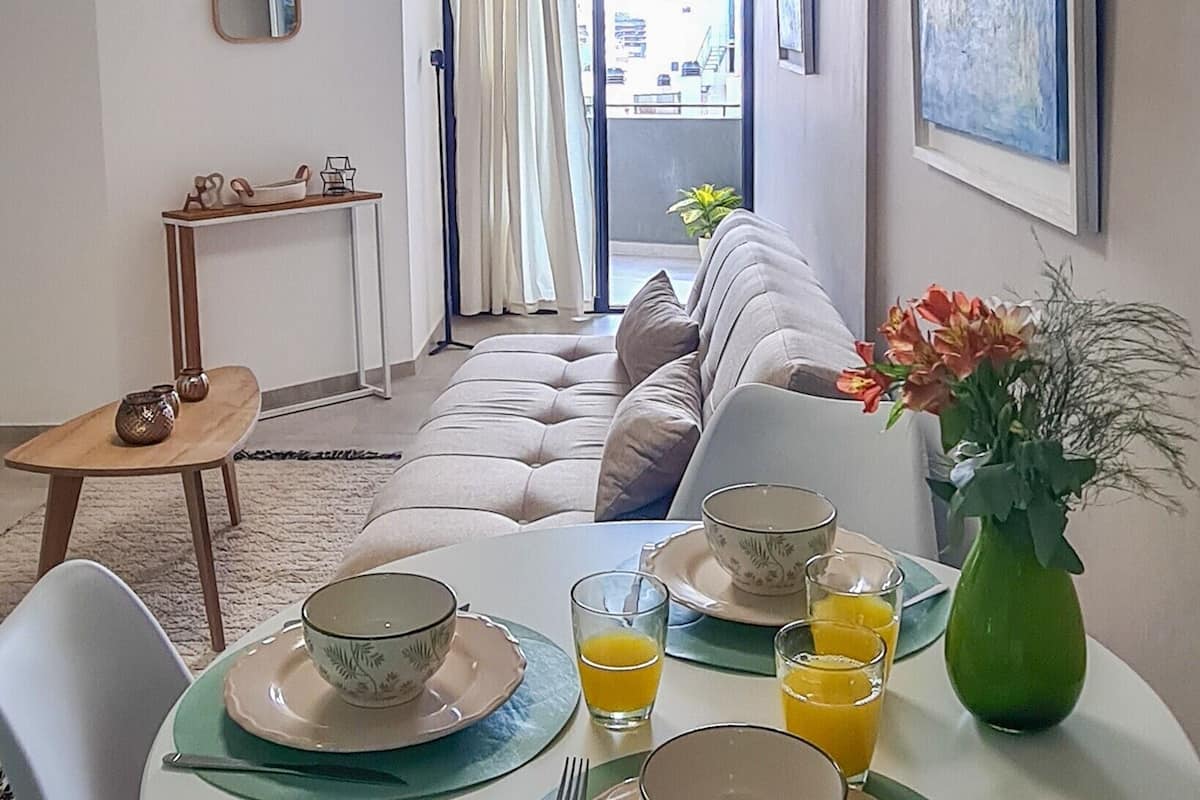 Uma sala de estar moderna com sofá, mesa de jantar posta com pratos, copos e um vaso de flores.