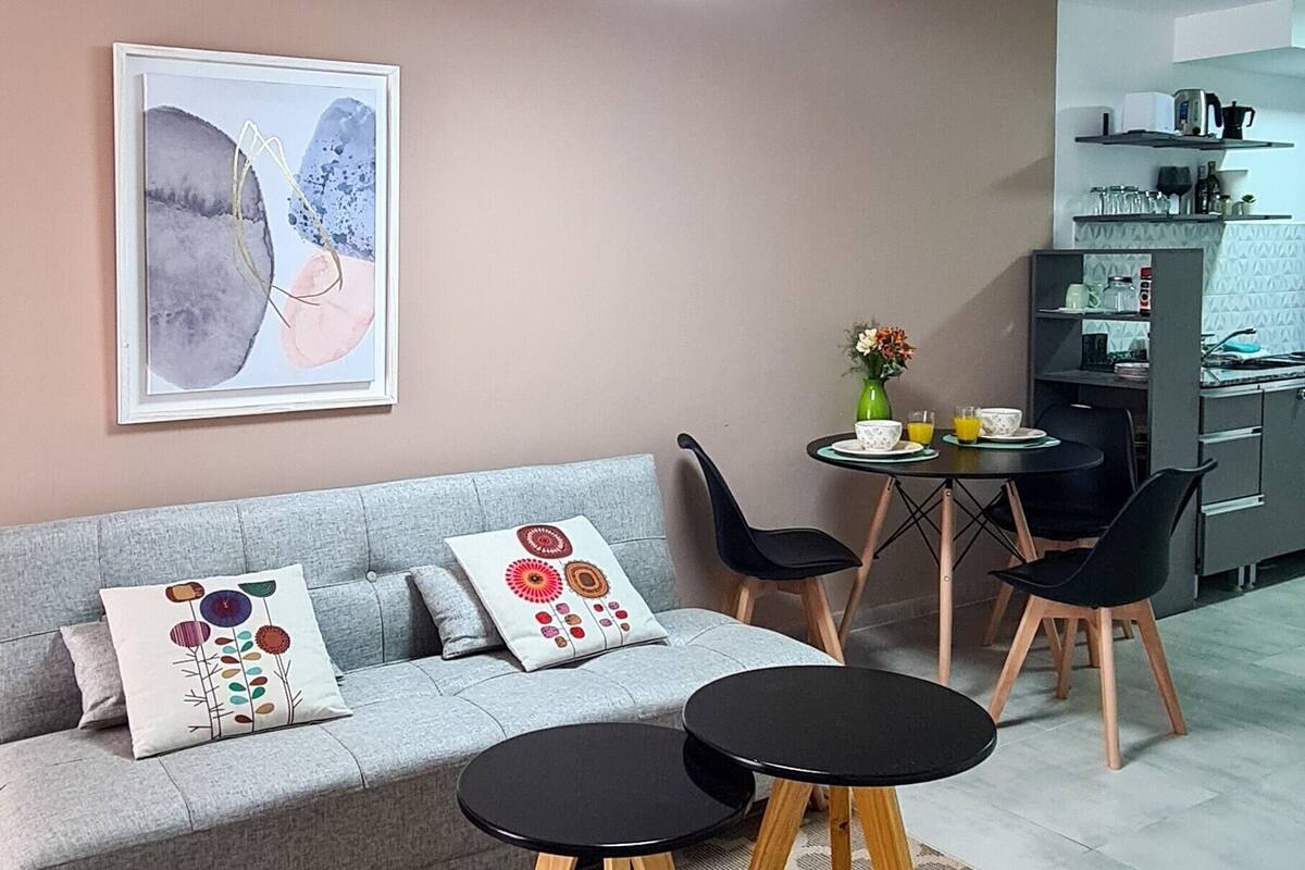 Uma sala de estar moderna com um sofá cinzento, duas mesas laterais pretas, uma mesa de centro e um espaço de jantar com uma mesa redonda pequena e cadeiras.