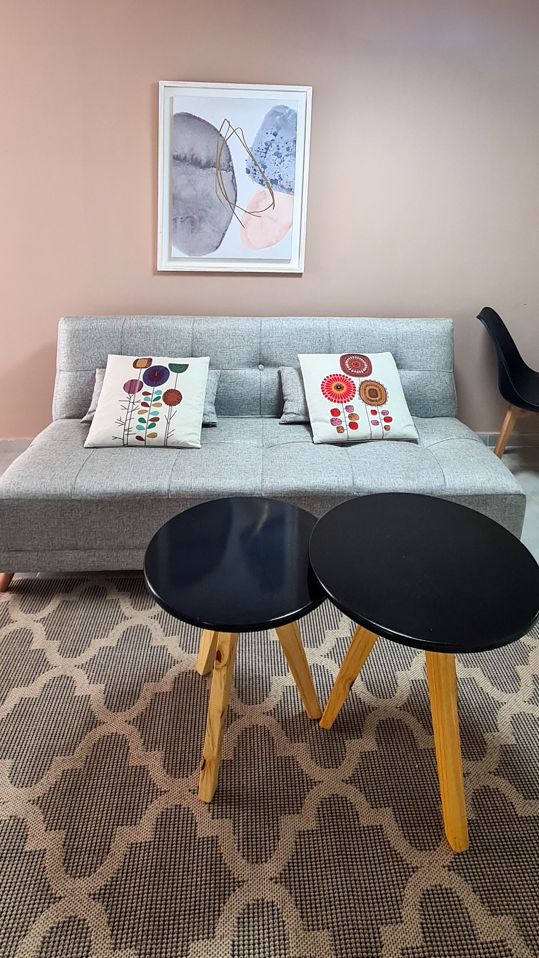 A grey sofa with floral cushions, a framed abstract painting, and a small black table with wooden legs.