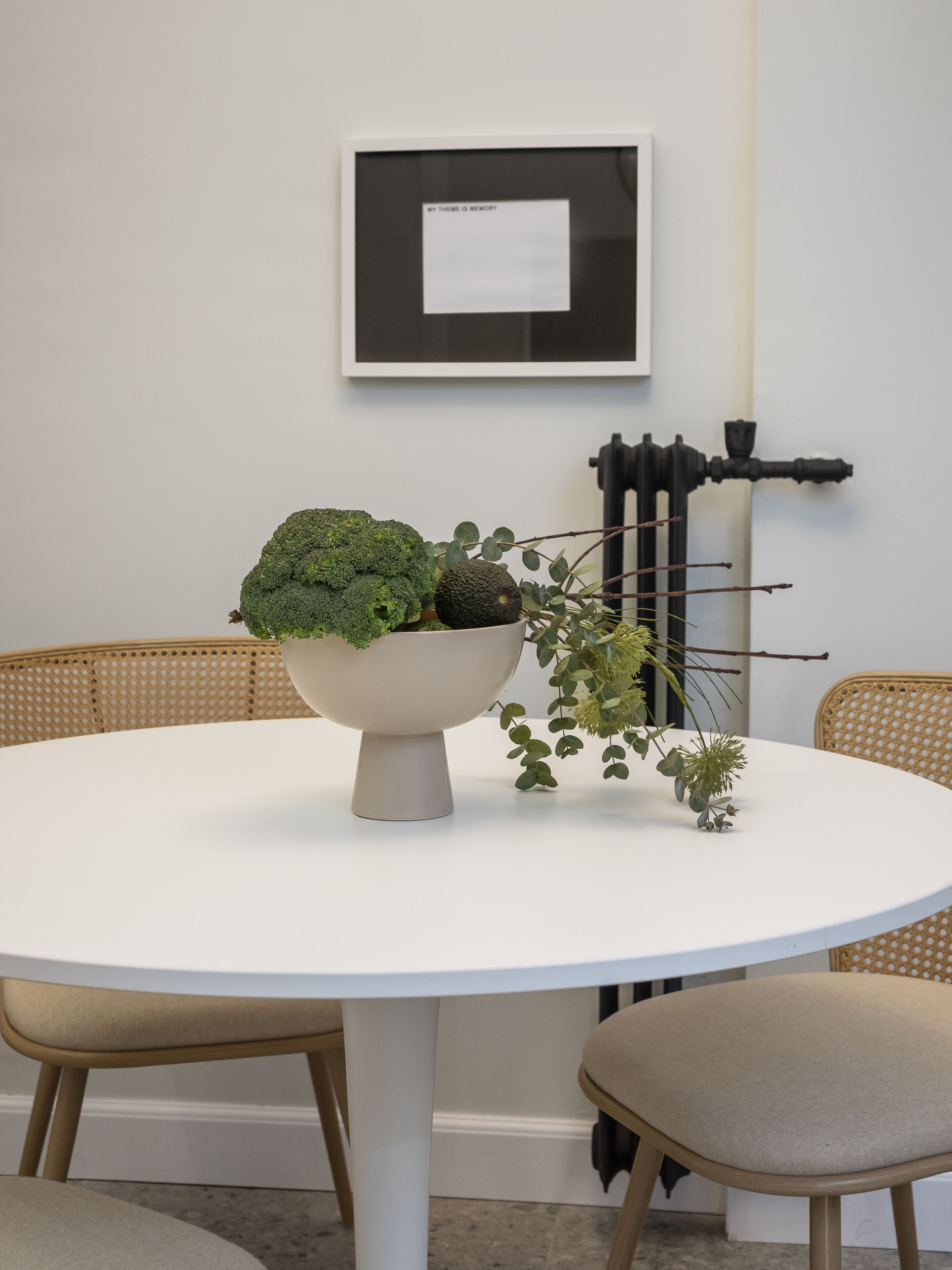 Une table ronde blanche, sur laquelle se trouve un saladier rempli de légumes verts et un pot de fleurs. Un tableau encadré est accroché au mur, au-dessus de la table.