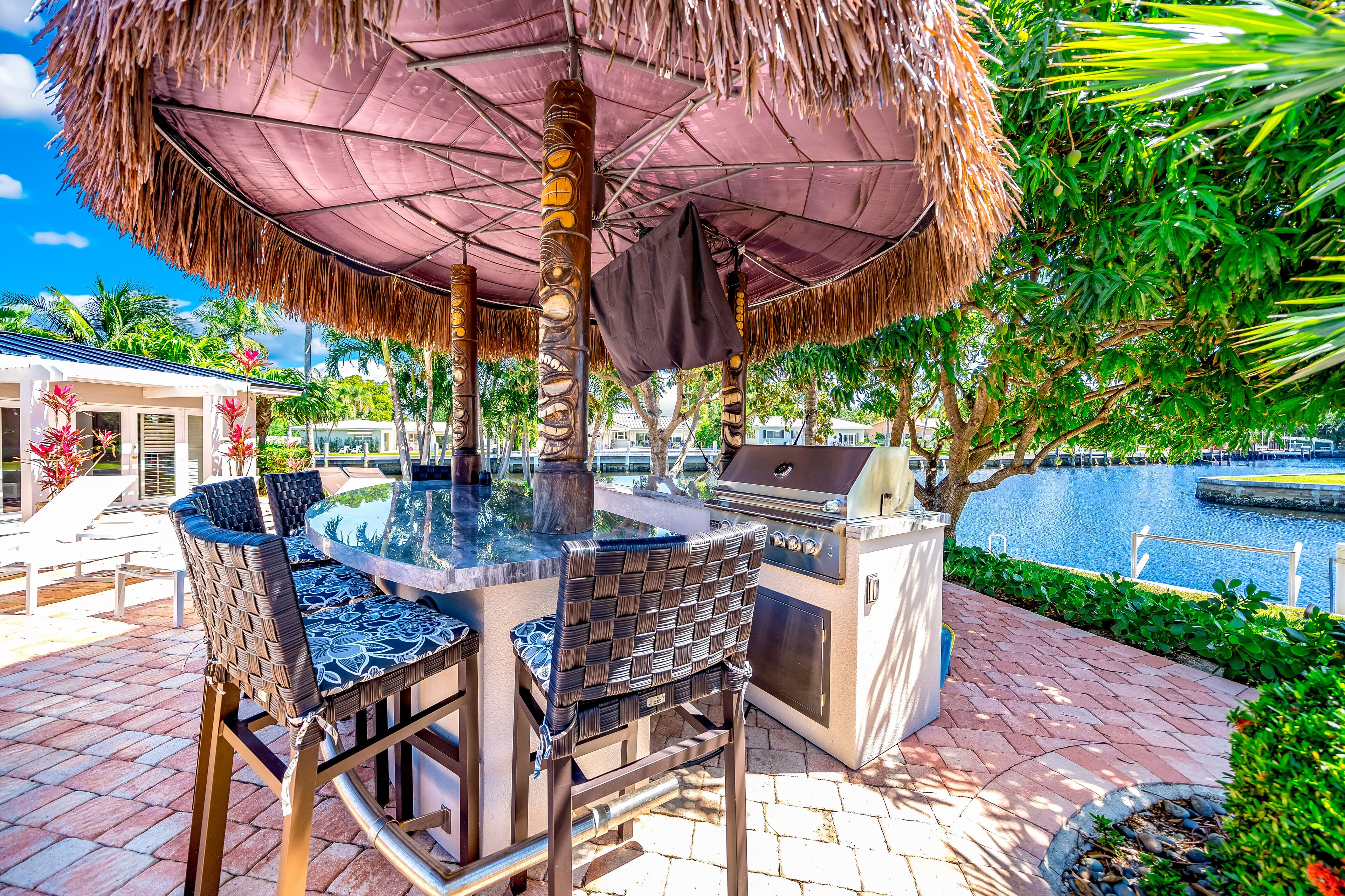 An outdoor kitchen area with a grill, dining table, and chairs under a thatched umbrella, surrounded by lush greenery and a water body.