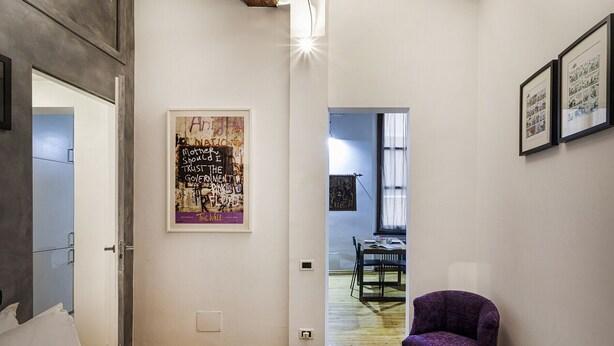 A hotel room with a bed, a purple chair, a poster on the wall, and a dining area visible through an open door. - Alle Mura del Molo Apartment by Wonderful Italy (Genova)