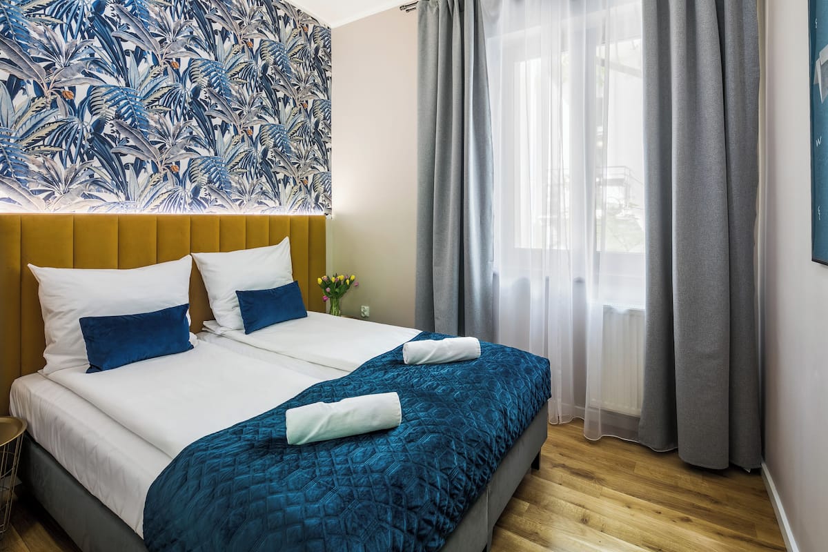 Une chambre d’hôtel avec un grand lit, une tête de lit, une fenêtre avec des rideaux et un mur orné d’un motif floral.