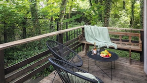 A wooden deck with a bench and two chairs, a table with a fruit bowl and a bottle, surrounded by lush greenery. - Garden House Apartments (Kraków)