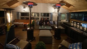 A cozy, well-lit living area with wooden floors and a high ceiling. There are two large, modern floor lamps, a comfortable seating area with sofas and cushions, and a small table.