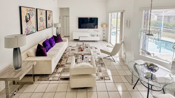 Een moderne woonkamer met een bank, salontafel en televisie. Er is een glazen tafel met stoelen en uitzicht op een zwembad.