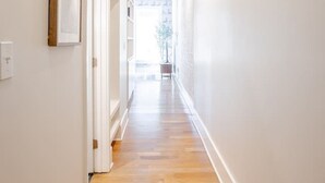 A bright, narrow hallway with wooden flooring and white walls, leading to a bright room with a large window.