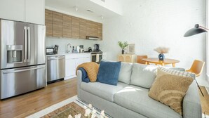 A modern kitchen with stainless steel appliances, a grey sofa with decorative pillows, and a dining area with a round table and chairs.