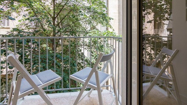 Un balcon avec deux chaises blanches et une rambarde en métal, d’où l’on peut admirer un arbre et des immeubles.