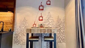 Ein gemütliches Zimmer mit Holzbalkendecke, Wandgemälde einer verschneiten Berglandschaft und einem Tisch mit Glasplatte.
