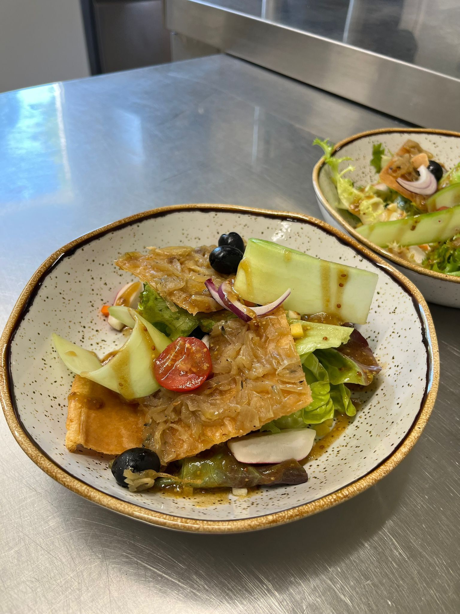 A bowl of salad with various vegetables and a piece of meat, served on a ceramic plate with a speckled design.
