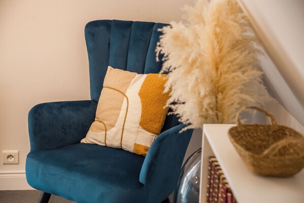 Un sillón azul con un cojín estampado, una planta decorativa y una cesta tejida sobre una mesa blanca.