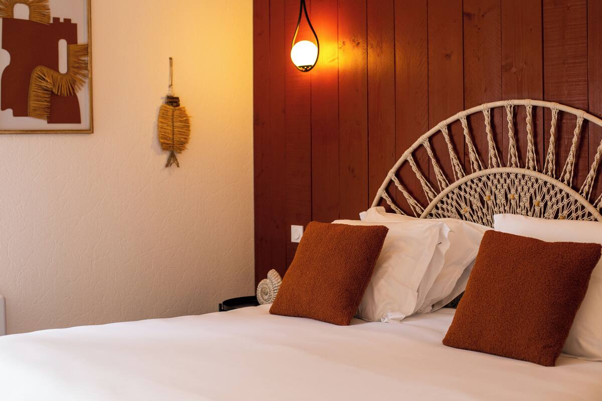 A bed with white linens and brown pillows, a woven headboard, a wall-mounted light fixture, and a wall decoration.
