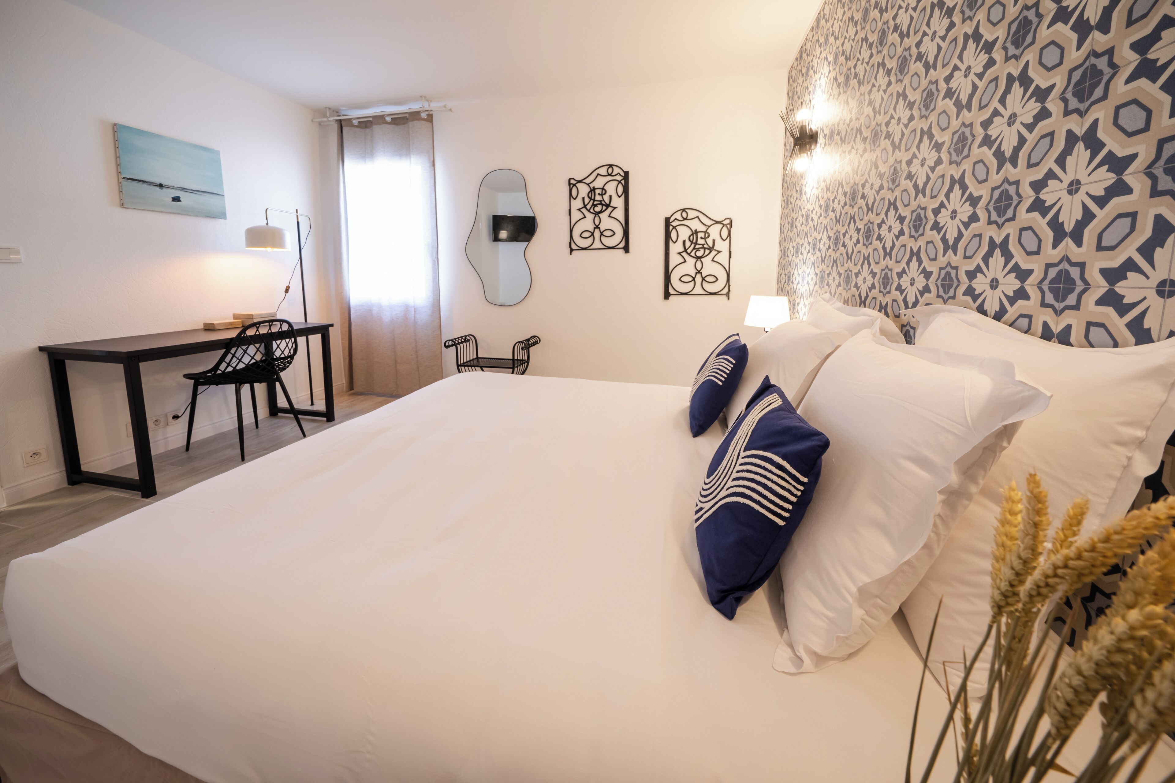 A bedroom with a large bed, a desk, and two chairs. There are three wall mirrors and a patterned wall.