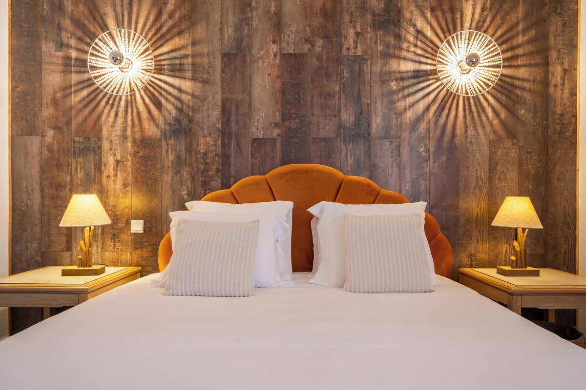 Een hotelkamer met een houten wand, een bekleed oranje hoofdeinde, witte kussens en twee bedlampen.