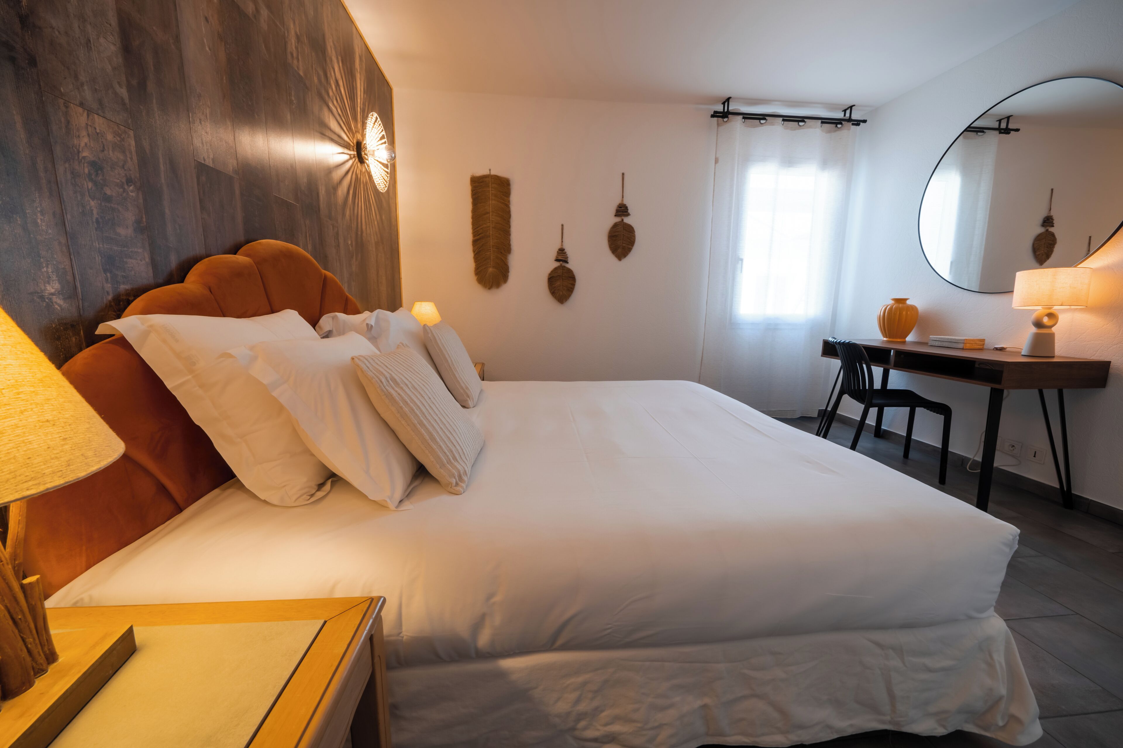 Une chambre avec un grand lit, un bureau et une chaise. Il y a une lampe sur le bureau et un miroir au mur.