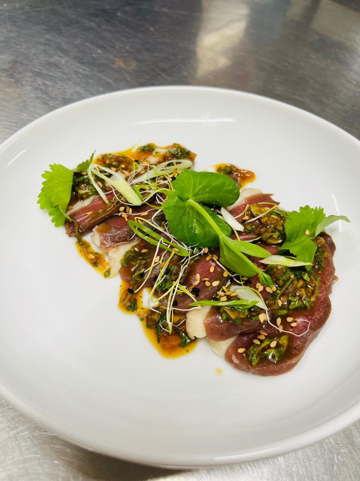 Une assiette blanche contenant une portion de viande coupée en fines tranches, des légumes verts et des jeunes pousses, le tout garni de graines de sésame et d’une sauce.
