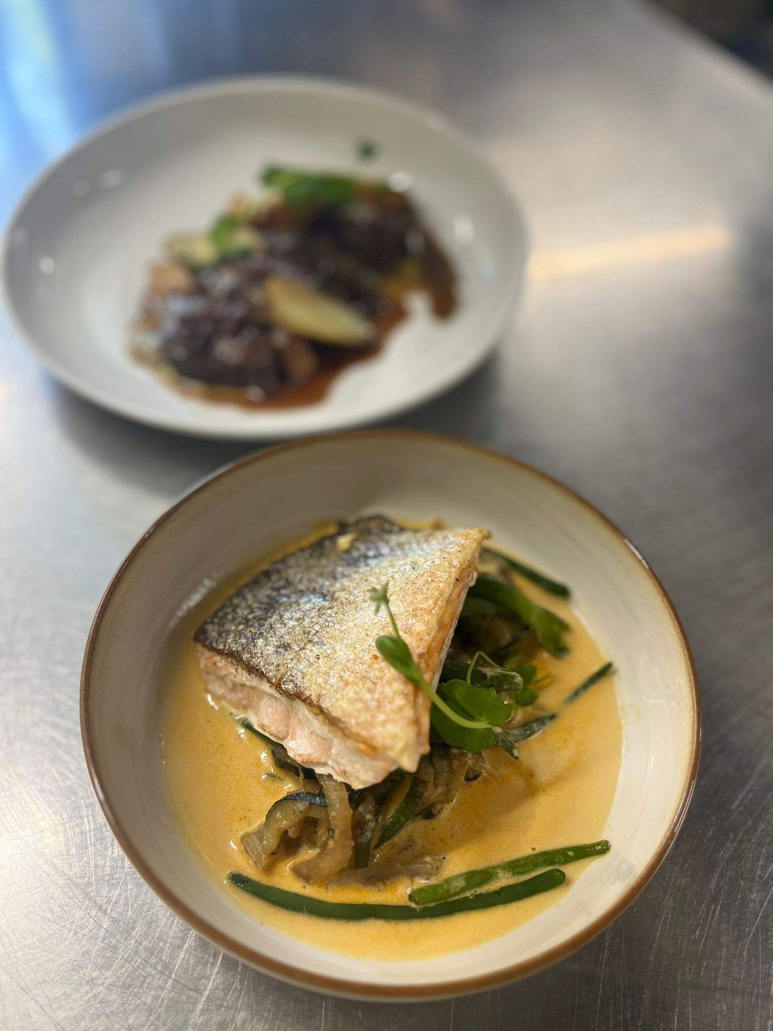 Ein Teller mit Fisch, grünem Gemüse und Soße, sowie ein weiterer Teller mit einem anderen Gericht im Hintergrund.