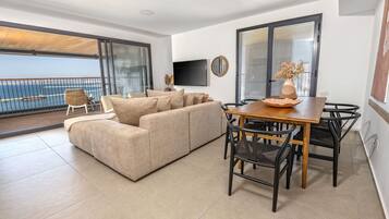 Un salon moderne comprenant un canapé, une table à manger, des chaises, un téléviseur et un balcon avec vue sur la mer.