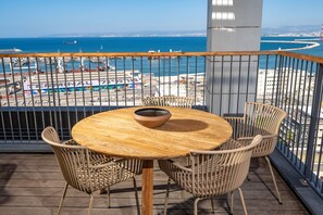 Mesa de madeira redonda com uma tigela, rodeada por cadeiras de vime, numa varanda com vista para um porto e edifícios.