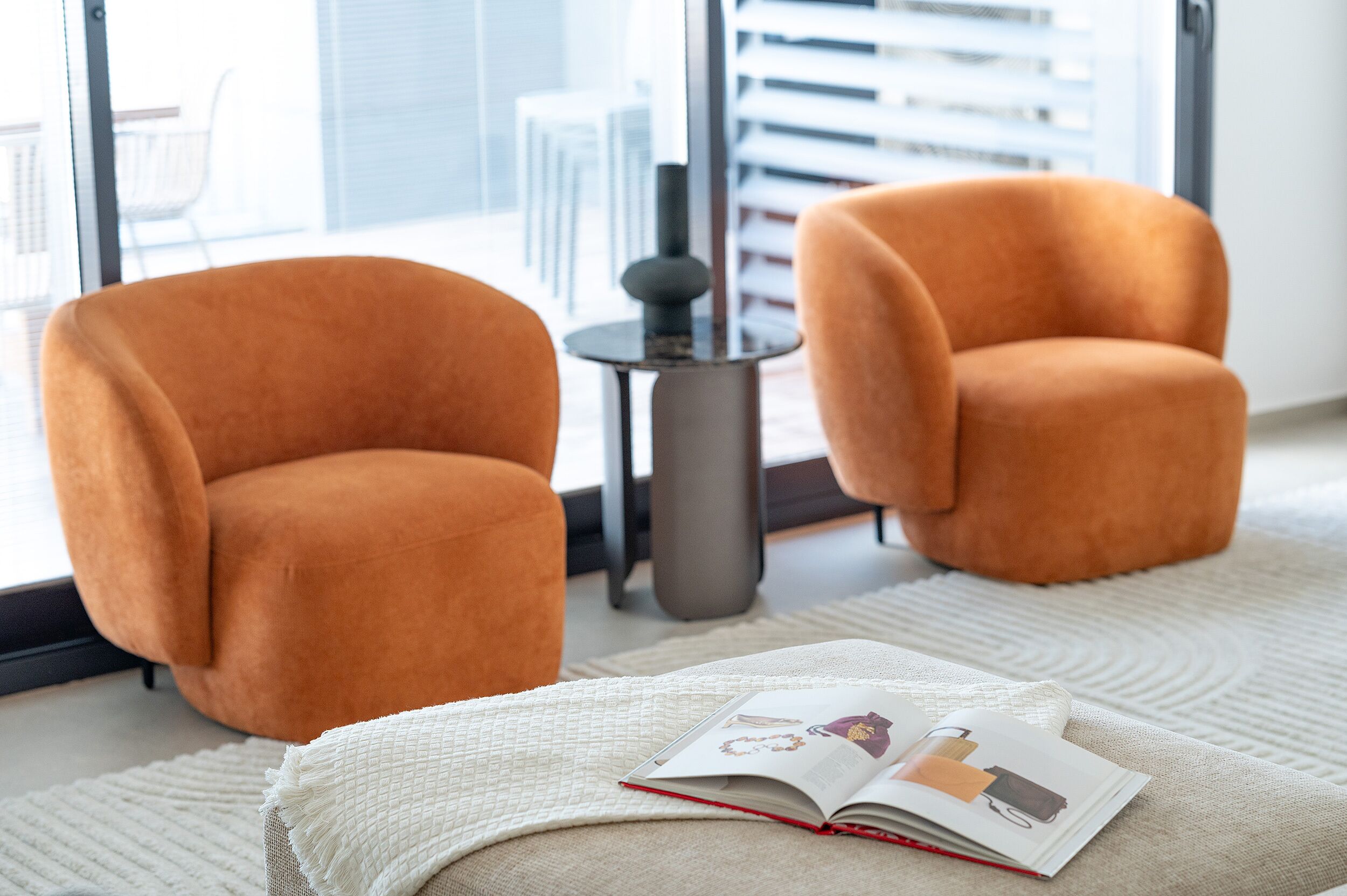 Two orange armchairs, a small round table with a lamp, and a book on a white textured rug in a modern room with large windows.