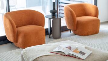 Two orange armchairs, a small round table with a lamp, and a book on a white textured rug in a modern room with large windows.
