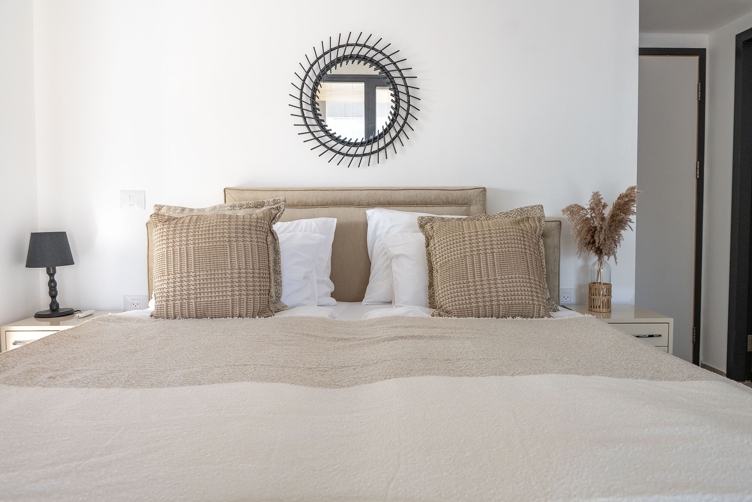 A neatly made bed with a beige blanket, two white pillows, and two textured pillows. A round mirror with a black frame hangs on the wall above the bed.