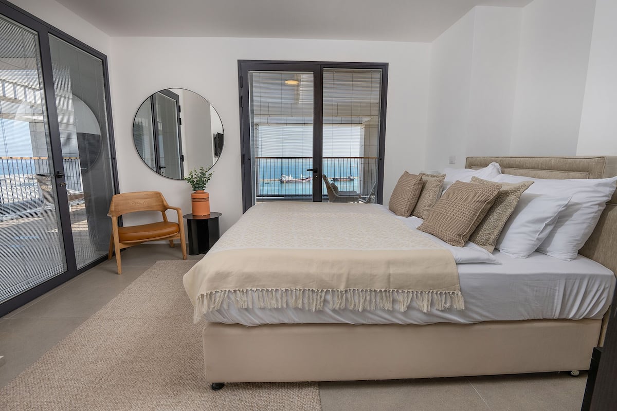 Un dormitorio con una cama grande, una silla, un espejo redondo y vista al mar a través de un ventanal corredera.