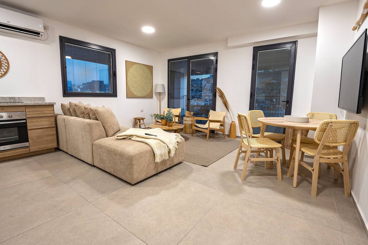 Una sala de estar moderna con sofá, mesa de comedor y sillas, zona de cocina con estufa y ventanas grandes con vistas a la ciudad.