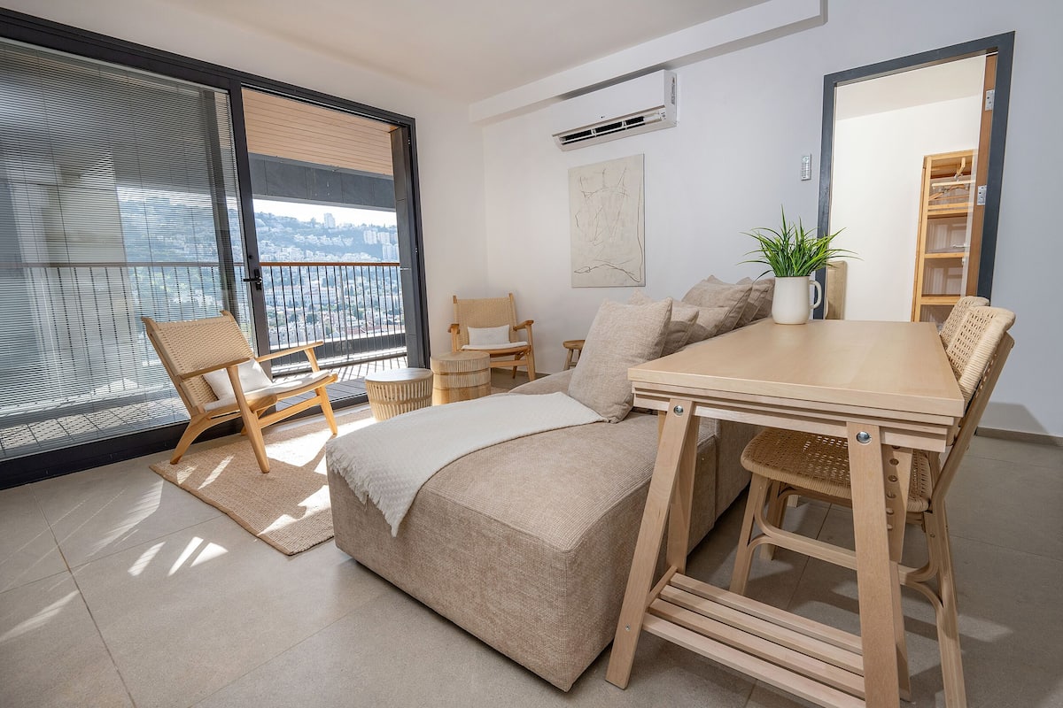 Un dormitorio moderno con un ventanal grande, una cama, una mesa de madera, una silla y una planta en maceta.