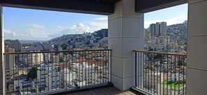 Varanda com grade de metal vista para a paisagem urbana, com vários edifícios e uma colina ao longe.