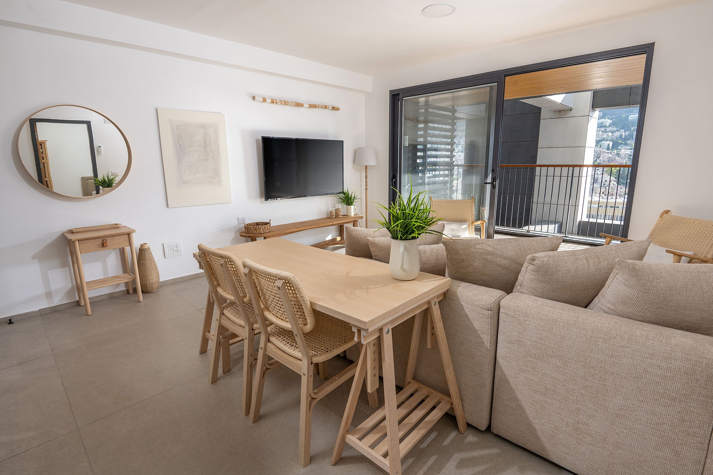 A modern living room with a wooden dining table, beige sofa, and a large window with a city view.