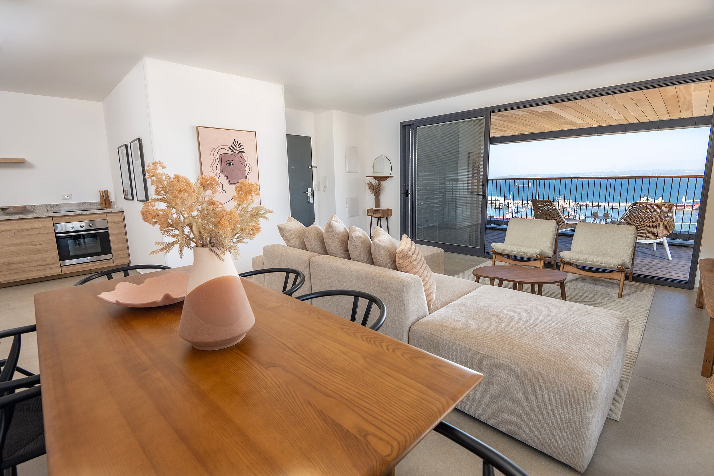 Una sala de estar moderna con una mesa de comedor de madera, un sofá beige y sillones, una zona de cocina y un balcón con vistas al mar.