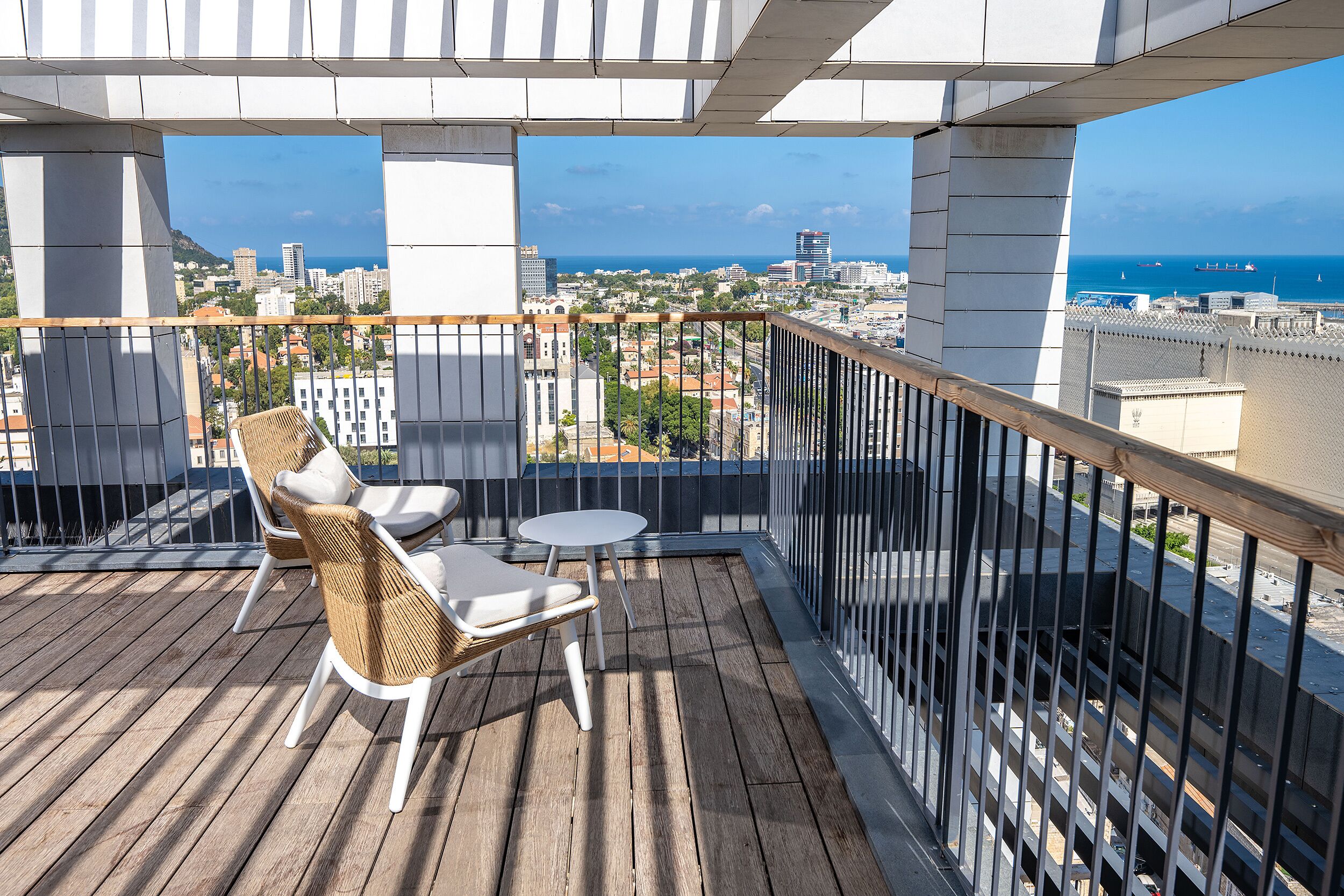 Ein Balkon mit Korbstühlen und einem kleinen Tisch, mit Blick auf eine Stadtlandschaft mit Gebäuden und dem Meer.