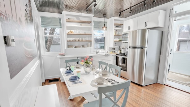 A modern kitchen with white cabinetry, a stainless steel refrigerator, and a dining area with a white table set for four. - Adorable Little Yellow House, Prime Location! Downtown Pella, IA (Pella)