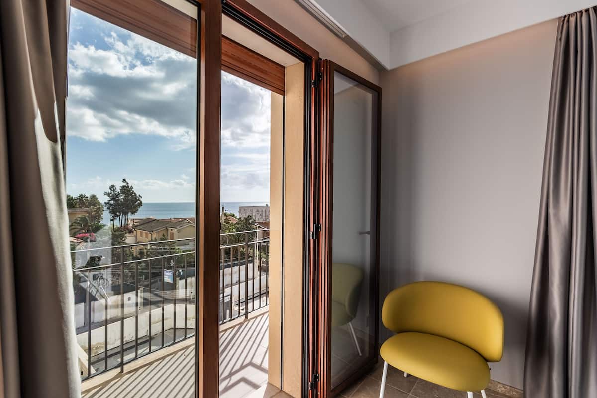 Un balcón con vista al mar y edificios, una silla amarilla y un ventanal corredera.