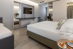 A modern hotel room with a large bed, a desk with a flat-screen TV, a chair, and a small table with a plate of food and a cup. - Hotel Netum Mare (Noto)