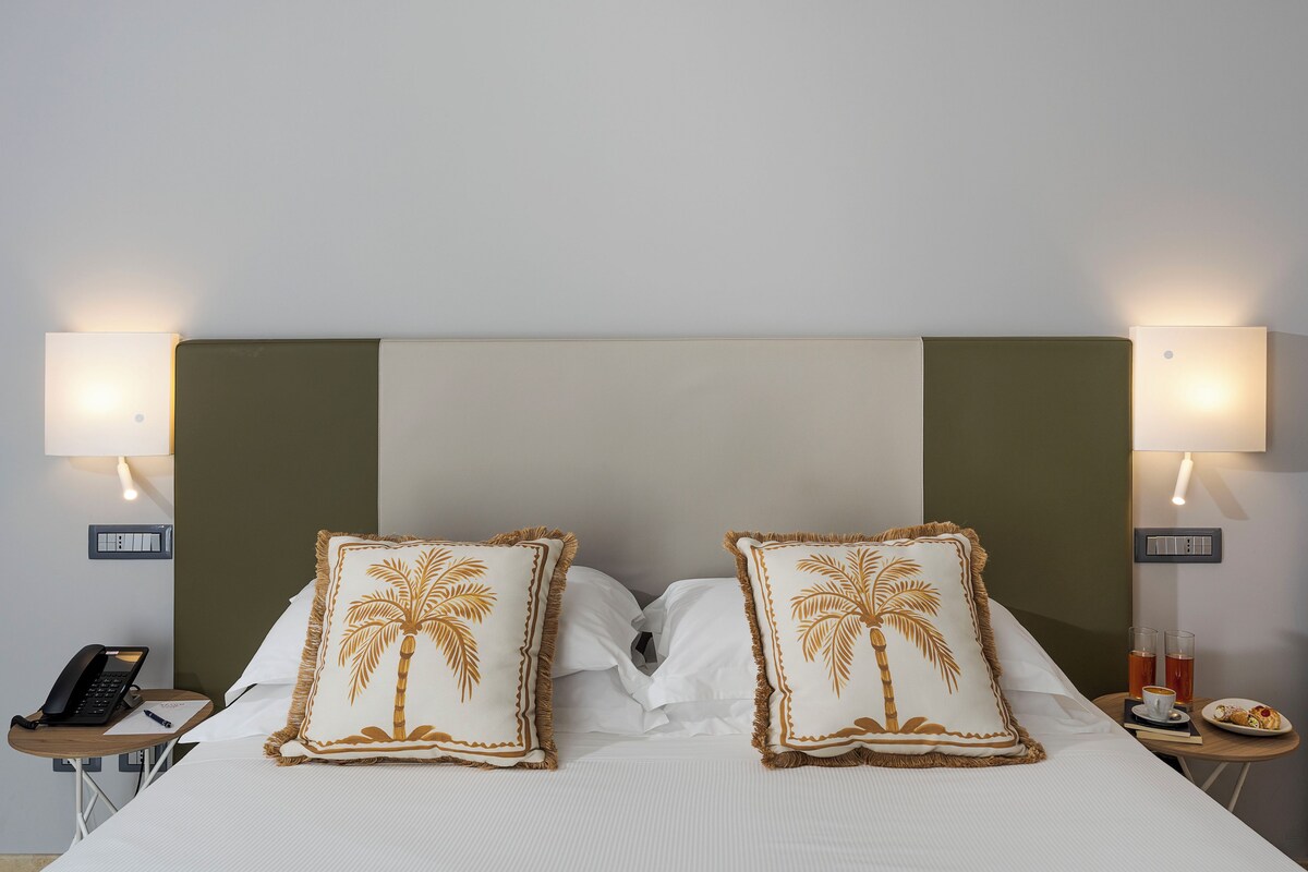 Habitación de hotel con cama, dos lámparas de noche, teléfono, una mesita con taza y pasteles, y un enchufe de pared.