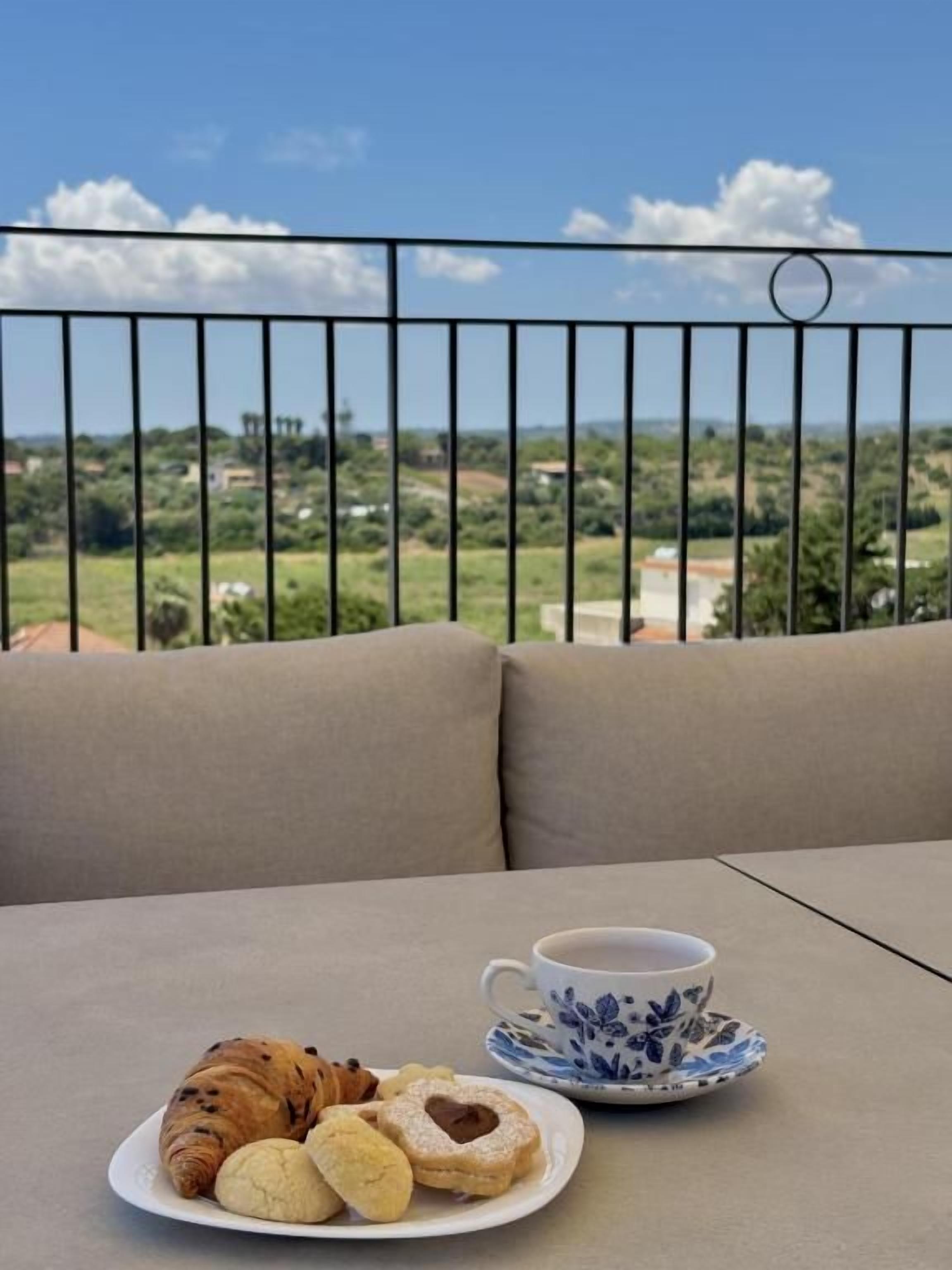 Un balcón con una mesa dispuesta para una comida, que incluye un croissant, un muffin y una taza de café, con una vista panorámica de fondo.