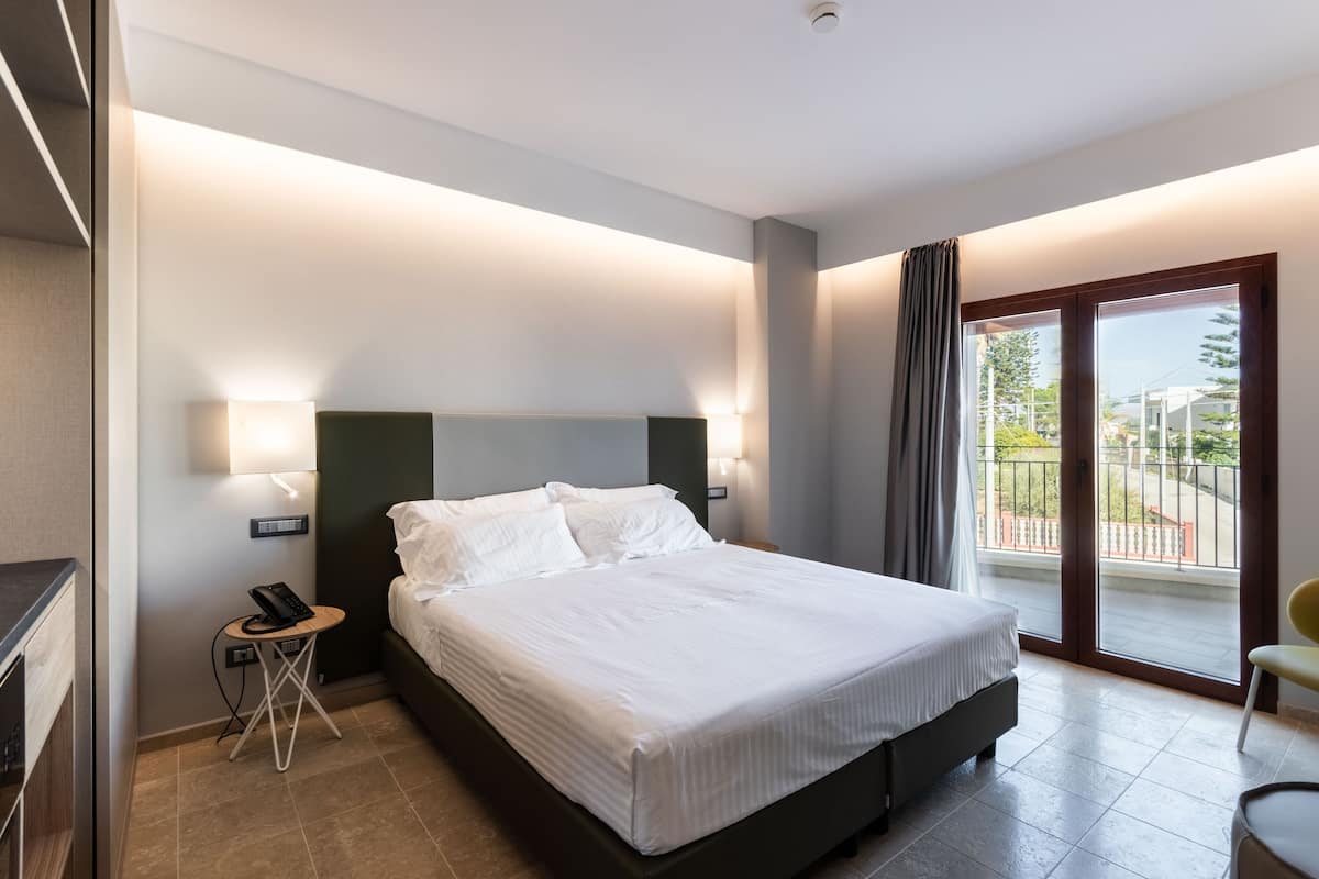 Habitación de hotel moderna con una cama grande, lámparas de noche, una mesita con un teléfono y vistas al balcón exterior.