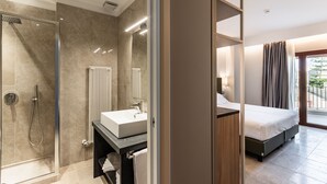 Habitación de hotel moderna con cabina de ducha de vidrio, lavamanos blanco, cama y vistas a un balcón.