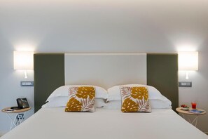 Una habitación de hotel con cama, dos lámparas de noche, una mesita con un teléfono y una mesita auxiliar con un vaso de jugo y un bol de fruta.