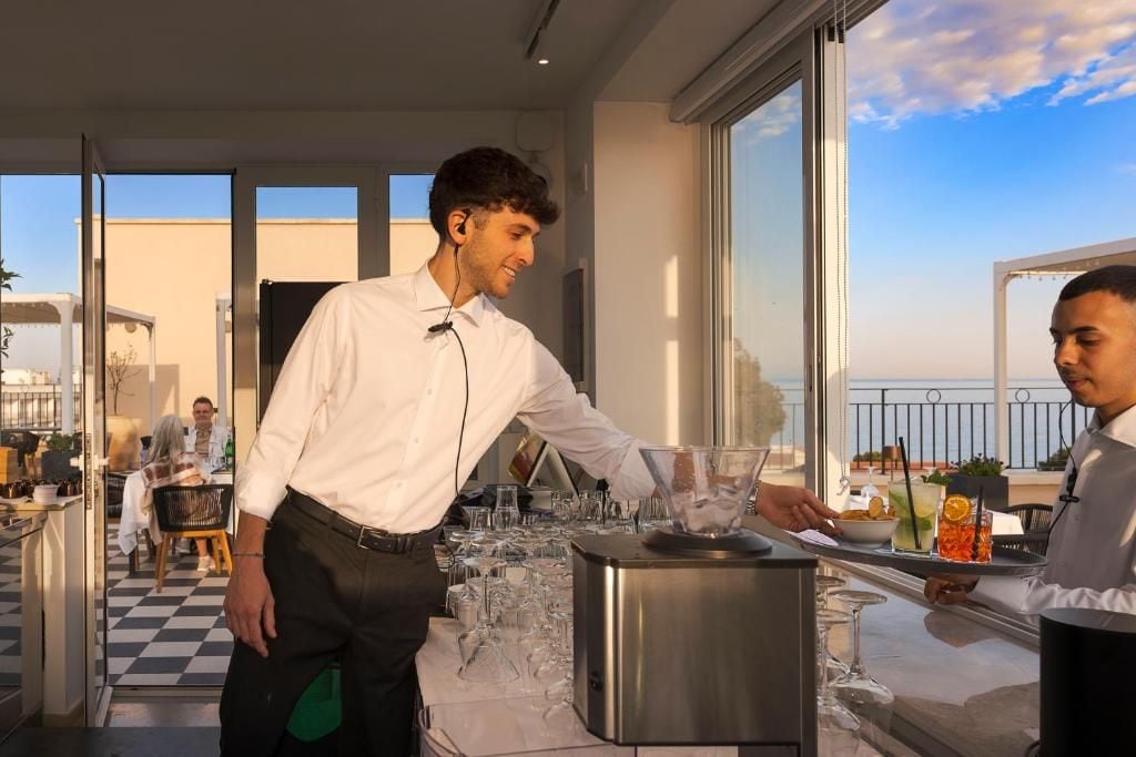 Un camarero sirve una bebida a un huésped en un moderno hotel con amplios ventanales y un balcón.