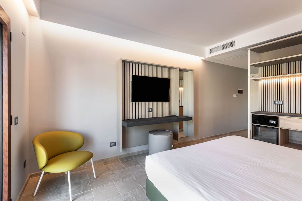 Habitación de hotel moderna con cama, televisor montado en la pared, zona de estar con sillón y escritorio integrado.